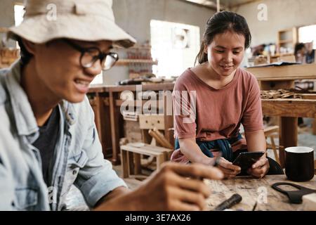 Asian female carpenter using smLifestylephone while sitting at workbench with her colleague in workshop Stock Photo