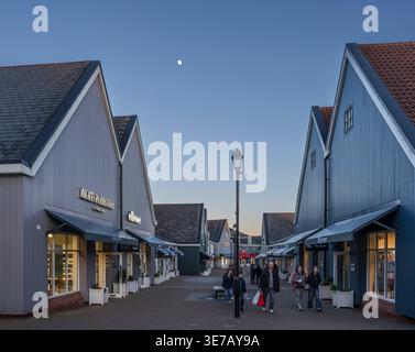 People stroll in the Caledonia Mall at Gretna lined with shops during a cool winter evening, illuminated by soft store lights and a distant moon Stock Photo
