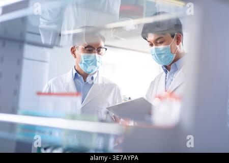 Chinese male researchers wearing lab gowns and face masks standing with clipboard working together in laboratory Stock Photo