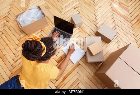 Black woman with braids using laptop taking note on notebook while lying on floor near box in living room at home Stock Photo