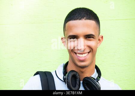 Smiling Arab male with headphones on neck standing against green wall during daytime Stock Photo