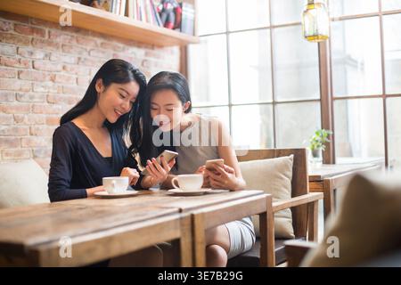 Smiling Chinese female friends sharing smartphone sitting at table with coffee cup in cafe during daytime Stock Photo