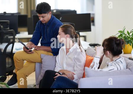 Middle Eastern man using tablet sitting on sofa beside caucasian female coworkers talking in office during break Stock Photo