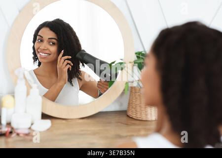 Black woman drying curly hair with hairdryer in front of mirror sitting at table in bedroom at home Stock Photo