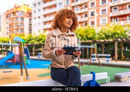 Mixed-race female teenager with blonde twists wearing brown jacket holding tablet and looking away while sitting on bench in city park during daytime Stock Photo