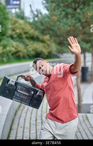 African American man holding boombox while dancing with raised hand in city park during daytime Stock Photo