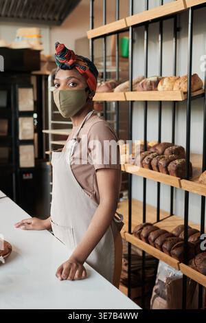Black saleswoman in headwrap wearing face mask and apron standing at table near bread racks in bakery shop Stock Photo