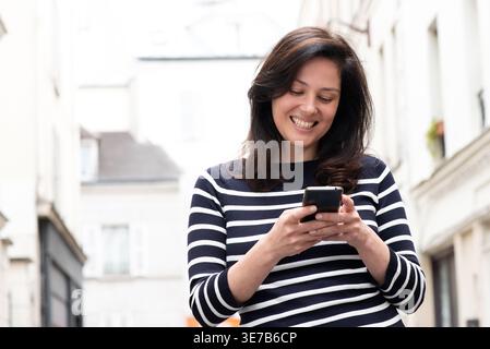 Smiling Asian woman using smartphone standing on street on sunny day Stock Photo