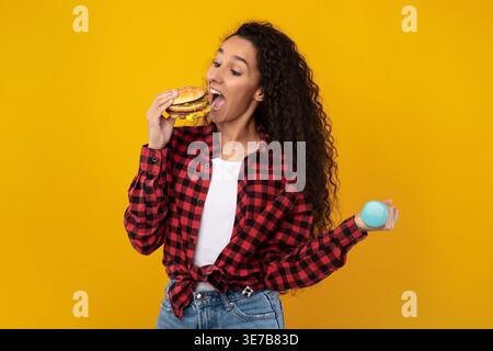 Middle Eastern woman with curly hair eating burger and holding dumbbell while standing against yellow background in studio Stock Photo