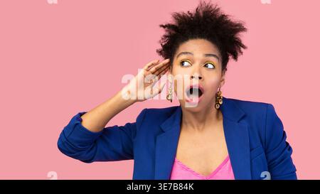 Black woman with open mouth holding hand near ear standing against pink background in studio Stock Photo