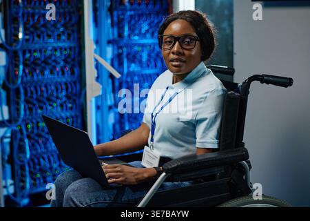 Mixed-race woman with eyeglasses using laptop while sitting on wheelchair in server room Stock Photo
