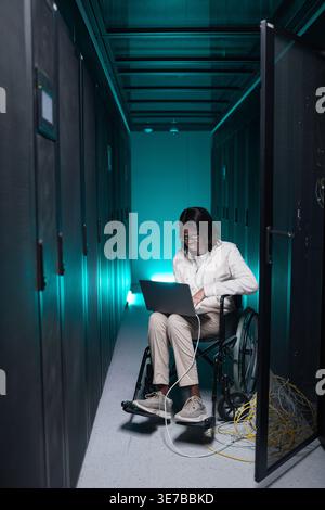 Disabled Black female IT engineer sitting in wheelchair wearing eyeglasses using laptop in server room Stock Photo