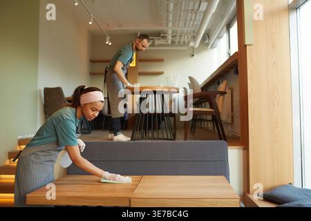 Mixed-race female and caucasian male employee in aprons and gloves wiping the tables in cafe Stock Photo