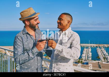 Smiling Mixed race and caucasian gay couple looking at each other drinking wine while standing on balcony against port with yachts in Mediterranean... Stock Photo