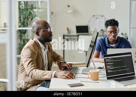 Serious Black male programmers sitting at desk developing new software on computers working together in office Stock Photo
