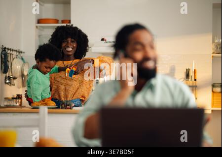 Cheerful Black mother with afro standing at kitchen counter having fun playing with her son while husband is working remotely at home Stock Photo