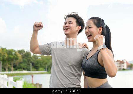 Cheerful Thai couple with raised arms and closed fists celebrating success after reaching their running target while standing together near lake in... Stock Photo