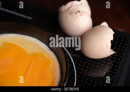close up of cracked eggshells and raw orange yolks in a cast iron skillet on a glossy black induction cooktop, warm rustic breakfast scene with soft m Stock Photo