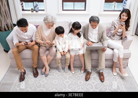 Smiling Chinese family using digital devices while sitting together on sofa at home Stock Photo