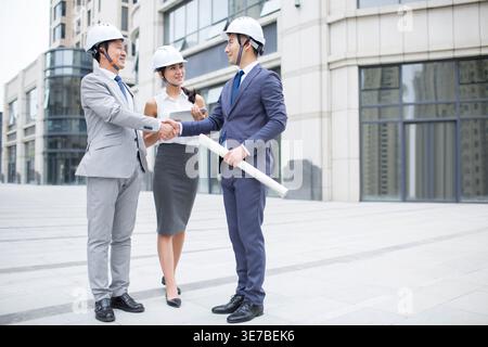 Smiling Chinese male and female architects wearing white hard hats shaking hands after closing business deal while standing on city street Stock Photo
