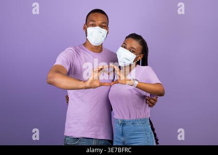 Black couple wearing medical face masks making heart shape with their hands and fingers while standing against purple background in studio Stock Photo