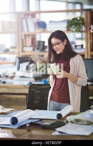 Chinese office worker wearing eyeglasses looking on her work and having cup of coffee while standing in office Stock Photo