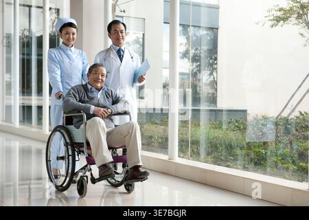 Smiling Chinese nurse pushing senior man sitting on wheelchair with doctor walking alongside in hospital corridor Stock Photo