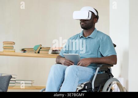Disabled Black man in VR goggles sitting in wheelchair using digital tablet for virtual therapy at home Stock Photo