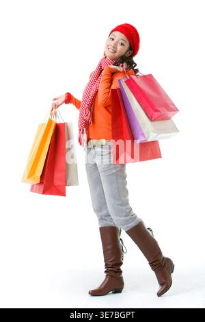 Smiling Chinese woman in beanie holding shopping bags while standing against white background in studio Stock Photo