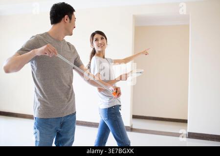 Smiling Chinese couple holding tape measure while standing at home Stock Photo