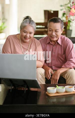 Asian senior couple having video call with family using laptop while sitting together on sofa at home Stock Photo
