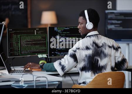 Black male programmer wearing headphones using laptop while sitting at table with computers in office Stock Photo