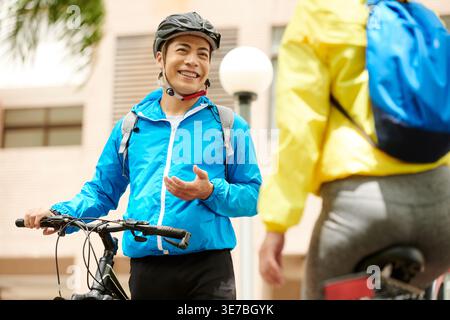 Smiling Asian man wearing blue jacket talking to girlfriend standing with bicycle on street Stock Photo