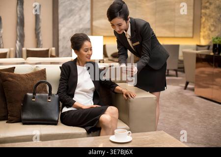 Chinese female staff showing menu to Chinese woman while sitting on sofa in hotel lobby Stock Photo