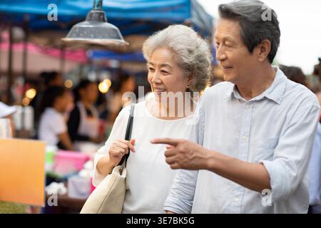 Smiling senior Chinese couple looking at street foods while walking together in street market during daytime Stock Photo