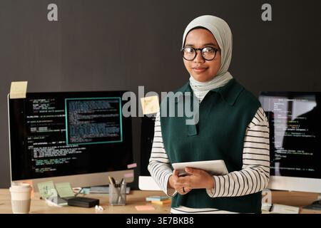 Mixed race Muslim female software developer wearing eyeglasses holding tablet standing against multiple computer screens placed on table in office Stock Photo