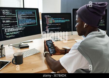 Black male software developer holding smLifestylephone listening to music with earphones sitting at desk with computer screens in workplace Stock Photo