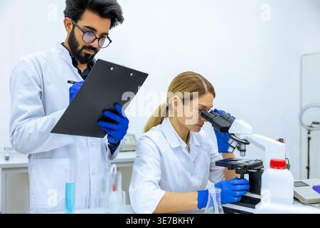Indian male scientist wearing lab coat and latex gloves making notes after doing sample test near caucasian female colleague using microscope in la... Stock Photo