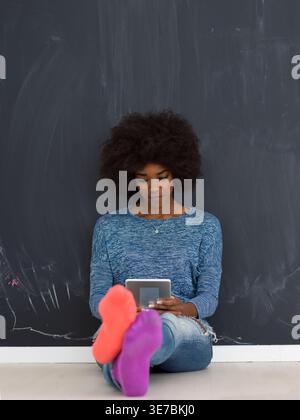 Focused Black woman with afro wearing colored socks using tablet sitting with crossed legs leaning against chalkboard wall in studio Stock Photo