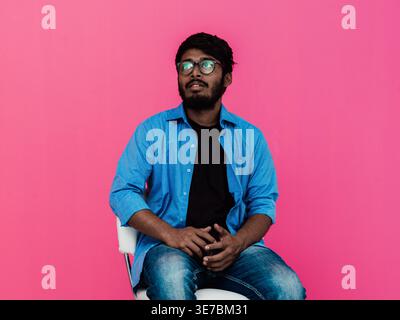 Serious Indian man in blue long sleeves with eyeglasses sitting on chair and looking away against pink background in studio Stock Photo