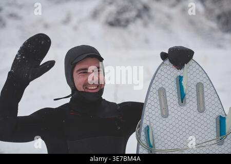 Smiling Mixed race man in surfing suit holding surf board and waving his hand while standing on snowy mountain during winter Stock Photo