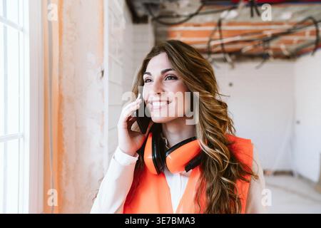 Smiling Hispanic female architect with headphones on neck wearing safety vest having phone conversation standing in construction site during daytime Stock Photo