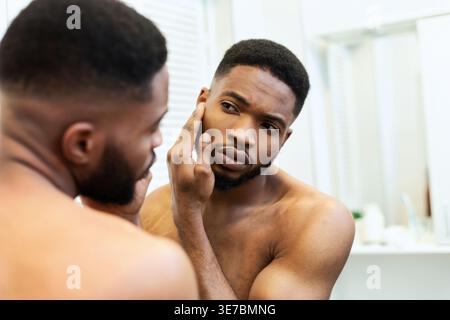 Serious Black man touching and checking his skin on face looking at mirror standing in bathroom at home Stock Photo