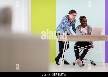 Black businessman with braids sitting at table working on laptop and having discussion with caucasian female colleague standing beside him in the o... Stock Photo