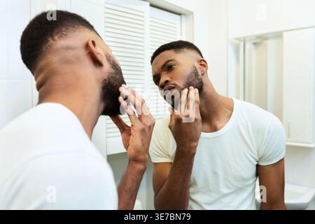 Worried Black man looking in mirror and checking his stubble standing in bathroom at home Stock Photo