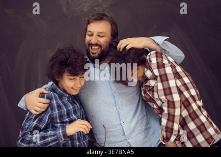 Cheerful Middle Eastern male teenagers in checkered long sleeves being hugged by caucasian man while standing against chalkboard background in studio Stock Photo