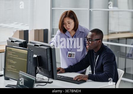 Asian female programmer in eyeglasses standing and pointing at monitor discussing computer program with Black male colleague in office Stock Photo