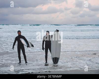 Smiling Mixed race men wearing surfing suits holding surfboards while standing on snowy beach during winter Stock Photo