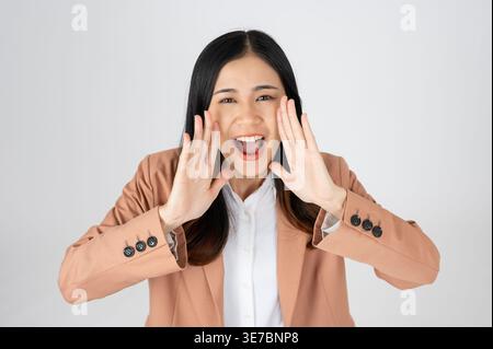 Smiling Asian female with hands near mouth screaming announcement standing against gray background in studio Stock Photo
