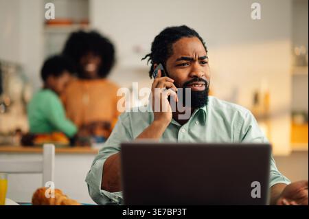 Serious Black father sitting at table with laptop having phone call working remotely near wife and son playing at kitchen counter at home Stock Photo